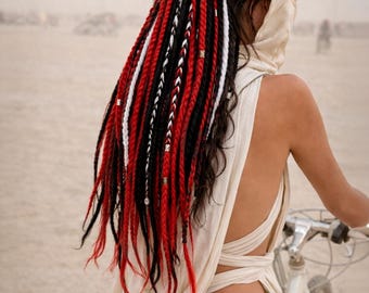 Op de afbeelding: Een persoon met donker haar in een knot, versierd met opvallende rode, zwarte en witte dreadlocks. Ze draagt een crèmekleurige hoodie en rijdt op een witte fiets in een woestijnachtige omgeving. De dreadlocks zijn versierd met kleine metalen kralen.