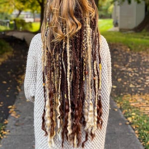 May include: Back view of a person with long, multi-toned hair styled in a half-up bun and adorned with dreadlocks. The dreadlocks are a mix of brown, blonde, and beige, with some strands featuring decorative beads. The person is wearing a light gray sweater.