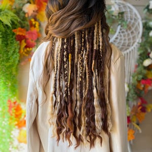 May include: A person with long, dark brown hair with blonde and brown dreadlocks. The dreadlocks are styled with gold accents. The person is wearing a beige blazer. The background features fall foliage.
