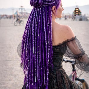 May include: A woman with long, vibrant purple dreadlocks, some accented with silver strands, styled in a bun and cascading down the back. She wears a dark, off-the-shoulder dress. The background shows a beach scene with people and bicycles.