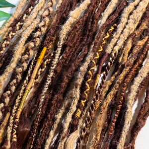 May include: Close-up of a collection of brown and blonde dreadlocks. The dreadlocks vary in thickness and texture, with some featuring intricate braids and wrapped accents. The color palette includes shades of brown, tan, and gold, creating a natural and textured appearance.