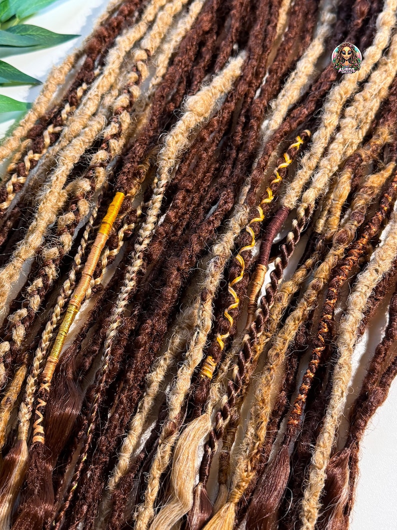May include: Close-up of a collection of brown and blonde dreadlocks. The dreadlocks vary in thickness and texture, with some featuring intricate braids and wrapped accents. The color palette includes shades of brown, tan, and gold, creating a natural and textured appearance.