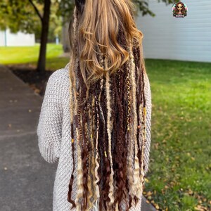 May include: A person with long, multi-toned hair extensions, featuring shades of brown, blonde, and beige. The extensions are styled with a mix of straight and curly textures, adorned with decorative beads. The person is wearing a light gray sweater.