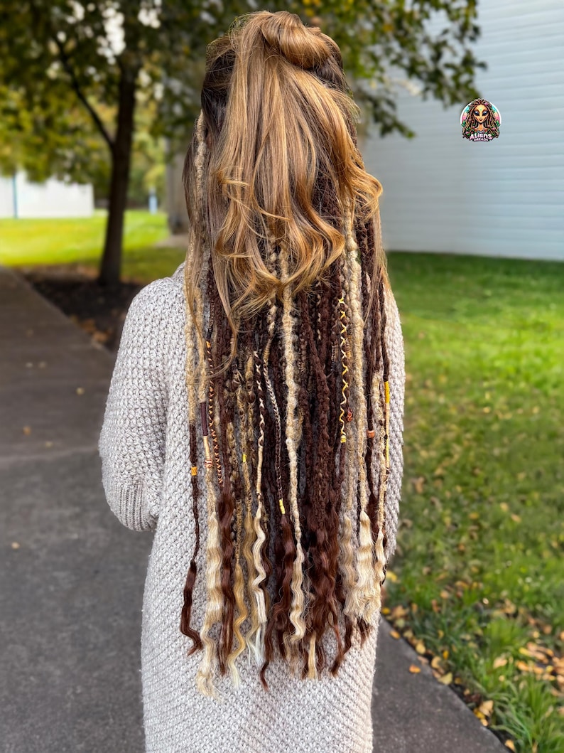 May include: A person with long, multi-toned hair extensions, featuring shades of brown, blonde, and beige. The extensions are styled with a mix of straight and curly textures, adorned with decorative beads. The person is wearing a light gray sweater.