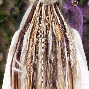 May include: A set of hair extensions featuring a mix of brown, blonde, and cream-colored dreadlocks and braids. The hair includes various textures, decorative wraps, small metallic beads, and light purple butterfly-shaped charms attached to strands.