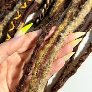 May include: Close-up of brown, blonde, and dark brown dreadlocks. The dreadlocks are a mix of colors and textures, with some having a twisted appearance. Some dreadlocks have yellow accents. The image is well-lit, showcasing the details of the hair.
