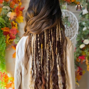 May include: A person's hair styled with a blend of dark brown and blonde dreadlocks. The hair transitions from dark to light brown, with blonde and gold accents. The person is wearing a beige blazer. The background features autumn foliage.