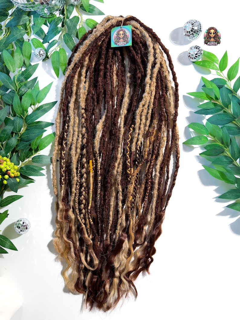 May include: A set of brown and blonde dreadlocks with wavy ends. The dreadlocks are a mix of dark brown, light brown, and blonde colors. A small tag with a logo is attached to the dreadlocks.