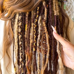 May include: A close-up of brown and blonde dreadlocks with yellow and brown accents. The dreadlocks are styled with various textures and patterns, including braids and wrapped sections. The hair is styled with a natural look.