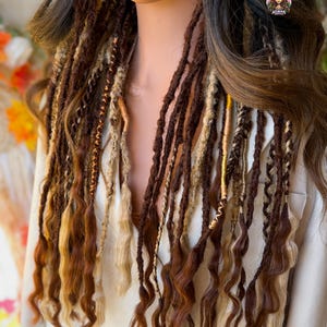 May include: A mannequin head displays long, wavy dreadlocks in shades of brown, blonde, and copper. The dreads are styled with gold and tan accents, creating a layered and textured look. The hair is arranged to cascade down the front, showcasing the color variations.