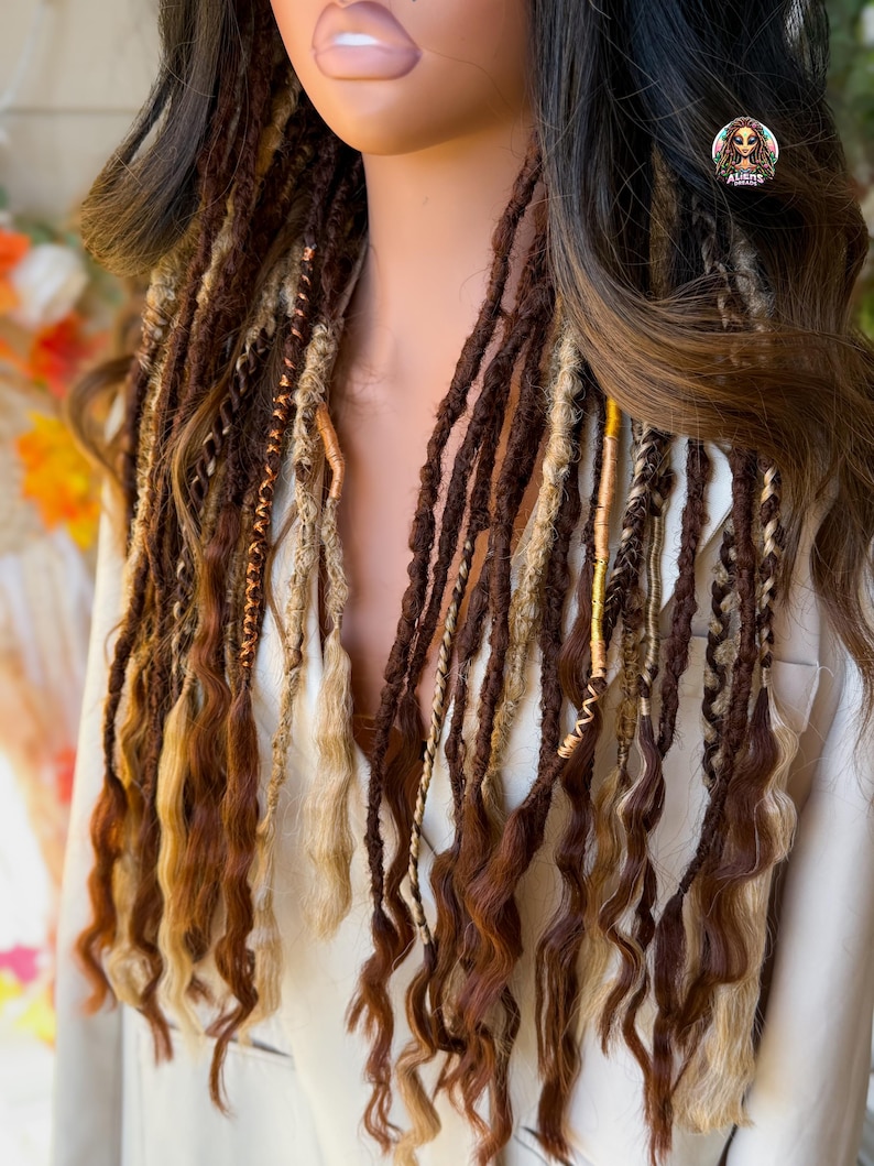 May include: A mannequin head displays long, wavy dreadlocks in shades of brown, blonde, and copper. The dreads are styled with gold and tan accents, creating a layered and textured look. The hair is arranged to cascade down the front, showcasing the color variations.