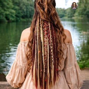 Puede incluir: Una mujer con cabello largo y ondulado castaño, peinado con múltiples trenzas en tonos rubios, rojos y marrones, adornadas con pequeñas cuentas. Lleva un vestido beige con hombros descubiertos, sentada junto a un lago.