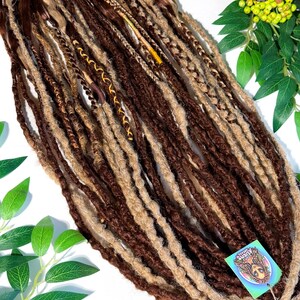 May include: A collection of brown and blonde dreadlocks, some with decorative wraps. The dreadlocks are arranged on a white surface with green foliage and a disco ball. A tag with a logo is attached to the dreadlocks.