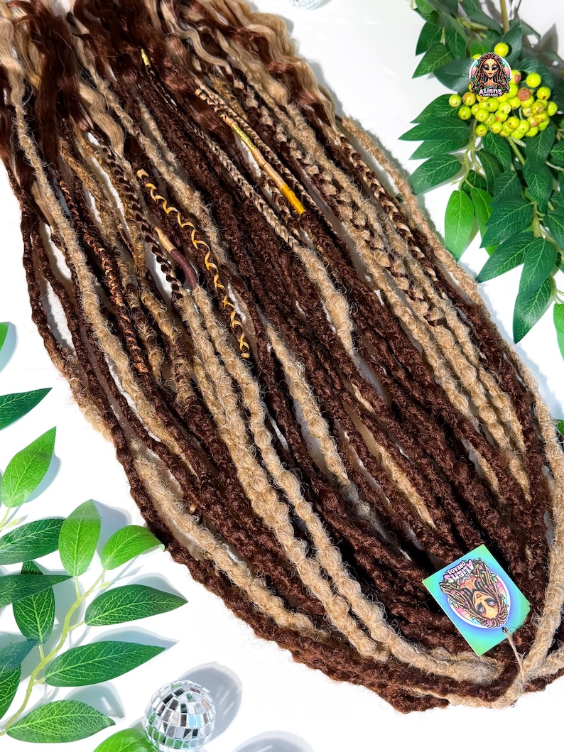 May include: A collection of brown and blonde dreadlocks, some with decorative wraps. The dreadlocks are arranged on a white surface with green foliage and a disco ball. A tag with a logo is attached to the dreadlocks.