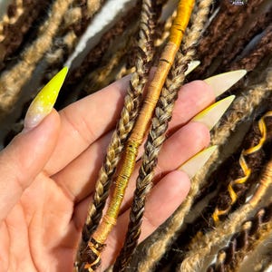 May include: Close-up of brown and blonde dreadlocks, some with gold accents. The dreadlocks are braided and wrapped with gold thread. The image shows a variety of textures and colors, with a focus on the intricate details of the hair.