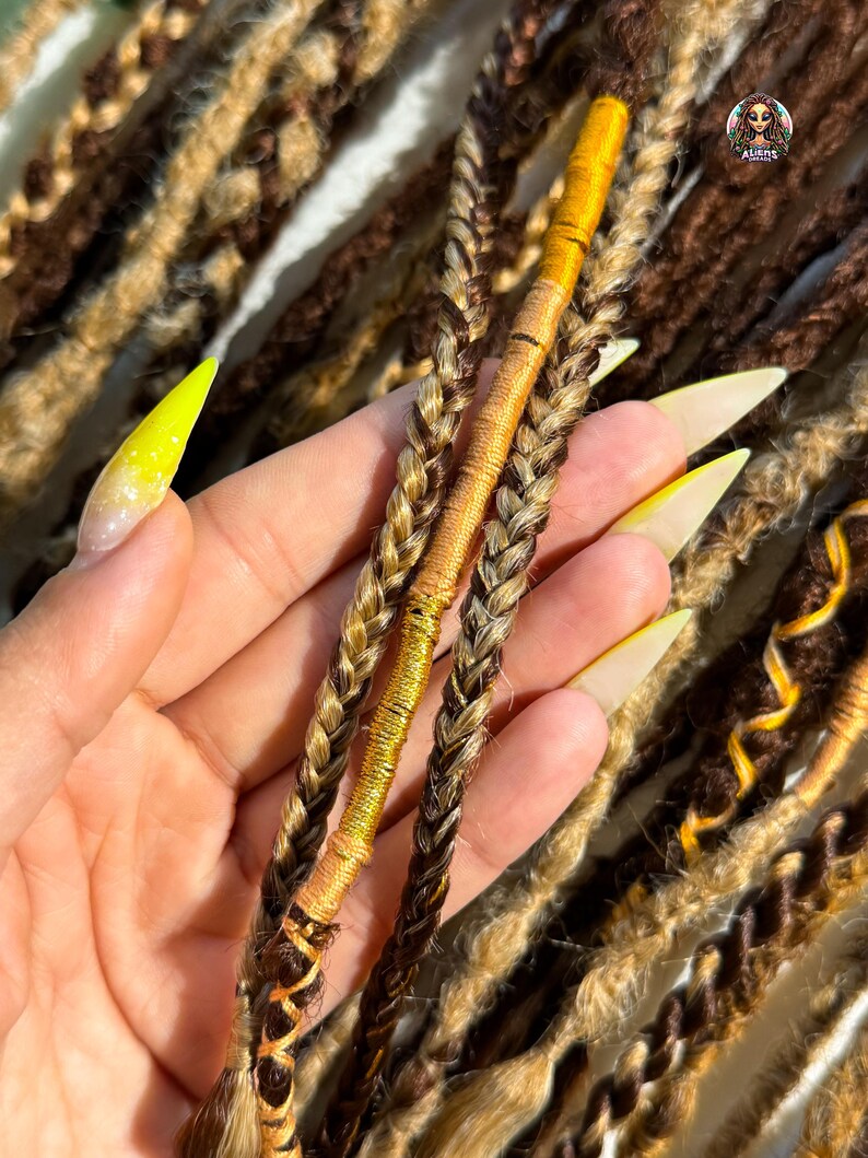 May include: Close-up of brown and blonde dreadlocks, some with gold accents. The dreadlocks are braided and wrapped with gold thread. The image shows a variety of textures and colors, with a focus on the intricate details of the hair.
