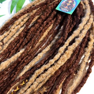 May include: A bundle of brown and blonde dreadlocks. The dreadlocks are a mix of dark brown and light blonde, with a textured appearance. A tag with the text "ALIENS DREADS" and an illustration of an alien is attached.