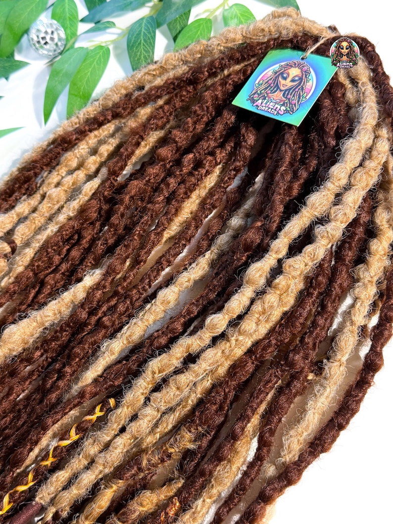 May include: A bundle of brown and blonde dreadlocks. The dreadlocks are a mix of dark brown and light blonde, with a textured appearance. A tag with the text "ALIENS DREADS" and an illustration of an alien is attached.