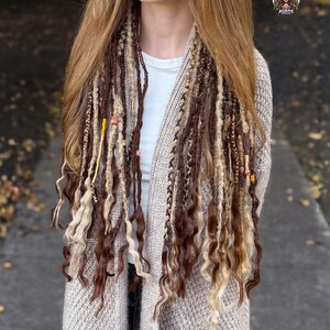 May include: A woman wearing brown and blonde dreadlocks with decorative beads. The dreadlocks are styled with a mix of textures and colors. She is wearing a beige cardigan over a white shirt.