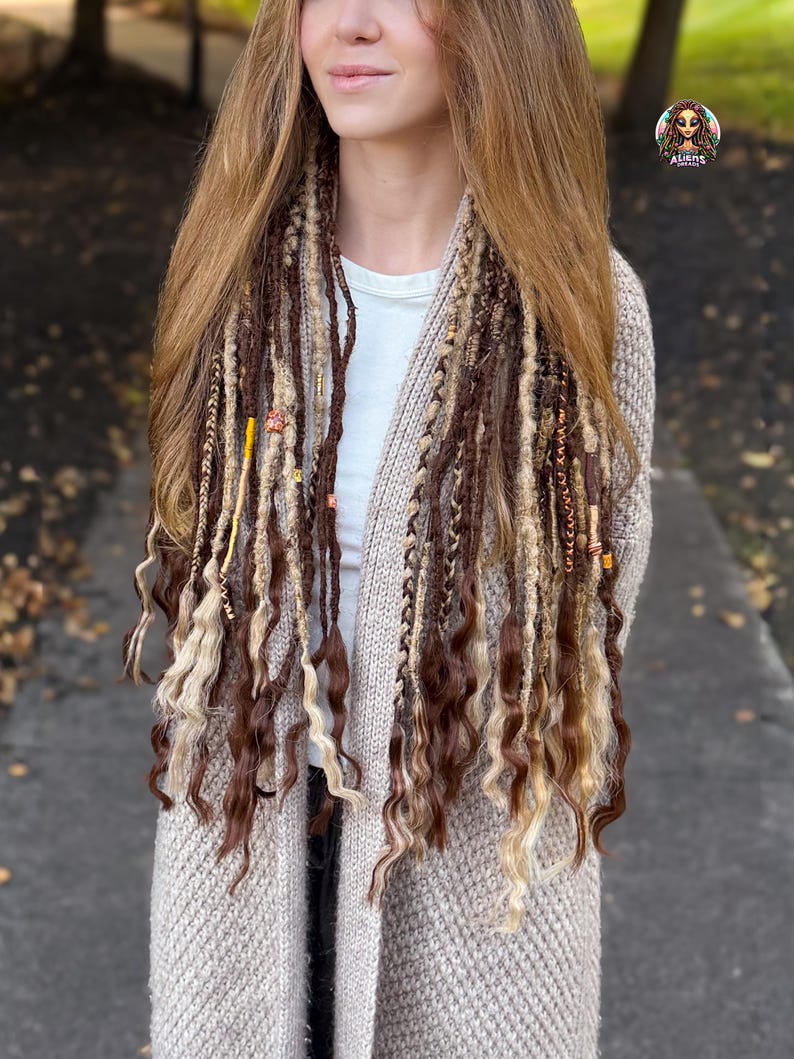 May include: A woman wearing brown and blonde dreadlocks with decorative beads. The dreadlocks are styled with a mix of textures and colors. She is wearing a beige cardigan over a white shirt.