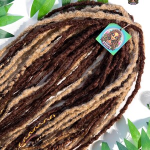 May include: A bundle of brown and light brown dreadlocks. The dreadlocks are arranged in a layered pattern, with a tag that reads "Aliens Dreads" attached. The background includes green leaves and a small disco ball.