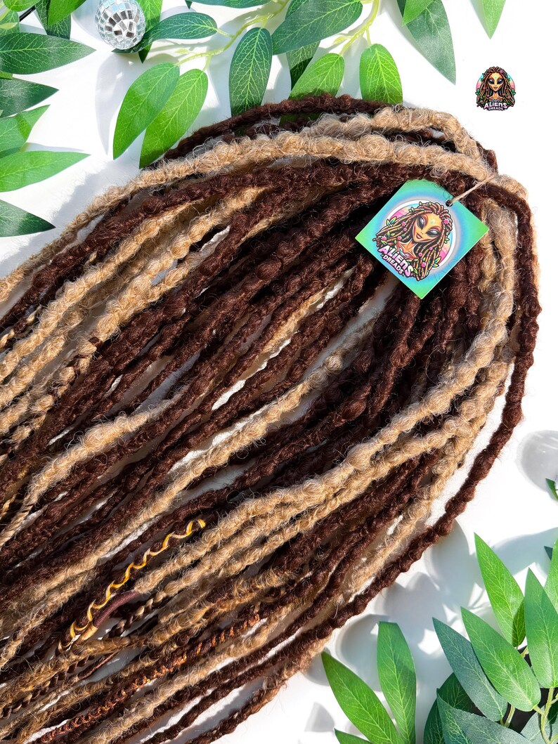 May include: A bundle of brown and light brown dreadlocks. The dreadlocks are arranged in a layered pattern, with a tag that reads "Aliens Dreads" attached. The background includes green leaves and a small disco ball.