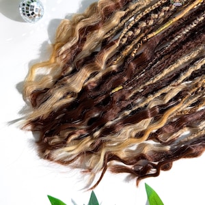 May include: A collection of brown, blonde, and light brown dreadlocks with wavy textures. The dreadlocks are arranged on a white surface, with a small disco ball and green leaves in the background. The dreadlocks have a logo that says "ALIENS".