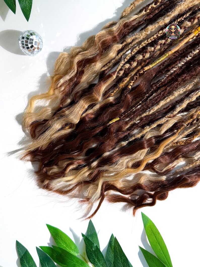 May include: A collection of brown, blonde, and light brown dreadlocks with wavy textures. The dreadlocks are arranged on a white surface, with a small disco ball and green leaves in the background. The dreadlocks have a logo that says "ALIENS".
