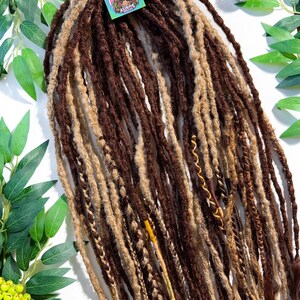 May include: A collection of brown and blonde dreadlocks, some with wavy ends, arranged on a white surface. A tag with a logo of a woman's face is attached. Green leaves and decorative mirrored balls are in the background.