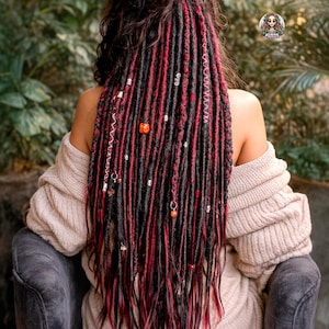 Puede incluir: Cabello largo y oscuro peinado con rastas negras y rojas, adornado con cuentas y abalorios decorativos. La persona lleva un suéter beige. El fondo presenta follaje verde.