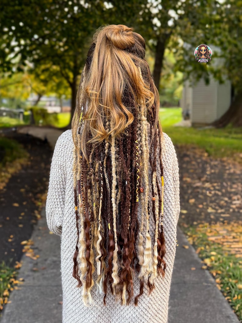 May include: Back view of a person with long, multi-toned hair styled in a half-up bun and adorned with dreadlocks. The dreadlocks are a mix of brown, blonde, and beige, with some strands featuring decorative beads. The person is wearing a light gray sweater.