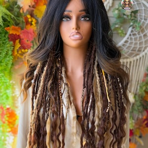 May include: A mannequin head with a black and brown ombre wig featuring long, wavy hair. The wig includes brown and blonde dreadlocks with gold accents. The background includes fall foliage and a macrame wall hanging.