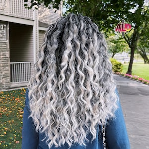 May include: A person with long, curly, black and white hair. The hair is styled in a loose, wavy pattern. The person is wearing a blue long-sleeved shirt.