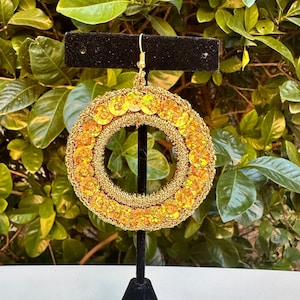 May include: Gold hoop earrings with iridescent sequins. The earrings are displayed on a black stand against a green leafy background.