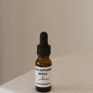 May include: A small amber glass bottle with a black dropper cap. The white label features black text that reads CAR DIFFUSER REFILL, Cashmere, and MILLER FARM CANDLE CO. The bottle is designed for dispensing liquid contents through the dropper top.