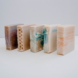 May include: Four bars of handmade soap in different colors and patterns. The bars are arranged in a row from left to right. The soap on the left is purple, the next is brown with a honeycomb pattern, the third is white with a blue swirl, and the last is beige with a light brown swirl.