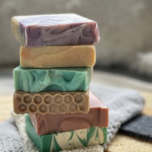 May include: A vertical stack of six rectangular bars of soap. The top bar is marbled purple, followed by a solid beige bar, a light green marbled bar, a tan bar with a honeycomb texture, a solid mauve bar, and a green and white marbled base bar.