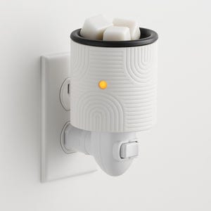 Peut inclure: Un diffuseur de cire &agrave; brancher blanc avec un bord noir et un motif g&eacute;om&eacute;trique. Le diffuseur contient des cubes de cire blancs et une lumi&egrave;re orange. Il est branch&eacute; dans une prise murale blanche.