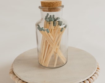 May include: A glass jar with a cork lid filled with wooden matches with blue tips. The jar is sitting on a white wooden pedestal with a beaded trim.
