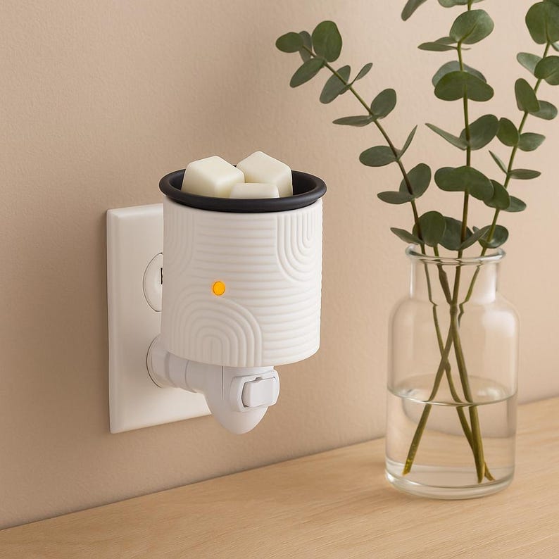 Peut inclure: Un diffuseur de cire &agrave; brancher blanc avec un dessus noir et un motif en arc d&eacute;coratif. Le diffuseur est branch&eacute; dans une prise murale et poss&egrave;de une petite lumi&egrave;re orange allum&eacute;e. Il contient des cubes de cire blancs. Un vase en verre transparent avec des tiges vertes est &agrave; droite.