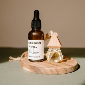 May include: A brown glass bottle with a black dropper top and a white label that reads "Car Diffuser Refill Blue Lava Miller Farm Candle Co.". The bottle is next to a wooden car diffuser with a clear glass container.