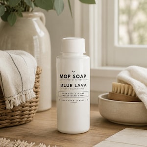 May include: A white bottle of "Mop Soap" floor cleaner concentrate, labeled "Blue Lava Fresh + Clean Citrus." The bottle is next to a woven basket with a towel, a ceramic vase with greenery, and a small bowl with brushes.