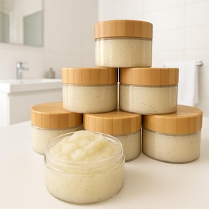 May include: A collection of small, clear glass jars filled with a light-colored, grainy substance, possibly a body scrub. The jars have natural wood-colored lids and are stacked on a white surface. The background is a bathroom setting.
