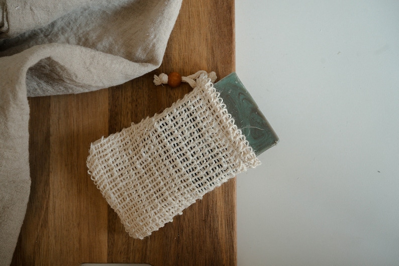 May include: A white mesh bag with a wooden bead drawstring, containing a green bar of soap.