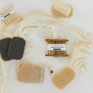 May include: A white cloth with two black loofahs, a brown bar of soap in a white mesh bag, a brown bar of soap in a wooden soap dish, and two brown loofahs. The soap dish is labeled "Bamboo Soap Dish Self Draining".
