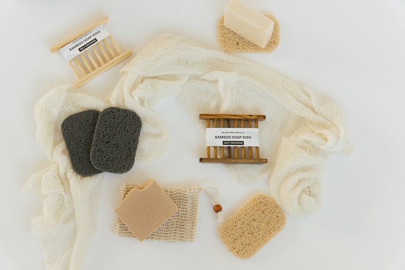 May include: A white cloth with two black loofahs, a brown bar of soap in a white mesh bag, a brown bar of soap in a wooden soap dish, and two brown loofahs. The soap dish is labeled "Bamboo Soap Dish Self Draining".