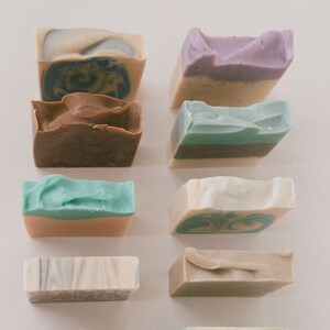 May include: Assortment of handmade soap bars in various colors and patterns, including brown, green, beige, and purple. The bars are arranged in two rows of five on a white surface.