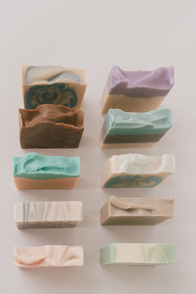 May include: Assortment of handmade soap bars in various colors and patterns, including brown, green, beige, and purple. The bars are arranged in two rows of five on a white surface.