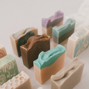 May include: Assortment of handmade soap bars in various colors and patterns, including brown, green, blue, purple, and white. The soaps are arranged in a row on a white surface.