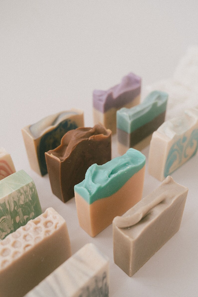 May include: Assortment of handmade soap bars in various colors and patterns, including brown, green, blue, purple, and white. The soaps are arranged in a row on a white surface.